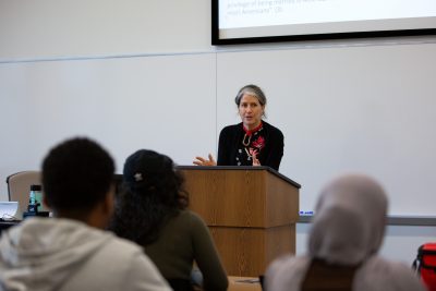 Kim is standing behind the lecturn. She is gesturing with her hands, fingers extended. Three students are visible in the foreground. They are not facing the carmera, they are facing Kim.