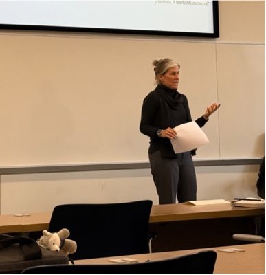 Professor Price-Glynn presenting in front of the class. she has her hair up, and is dressed in a black sweater and grey pants, holding lecture slides.